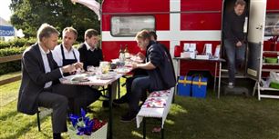 Dansk Folkepartis Dannebro-campingvogn er kørt i stilling på Bornholm. Uden for sidder Kristian Thulesen Dahl og Peter Skaarup og spiser flæskesteg og brun sovs i aftensolen.