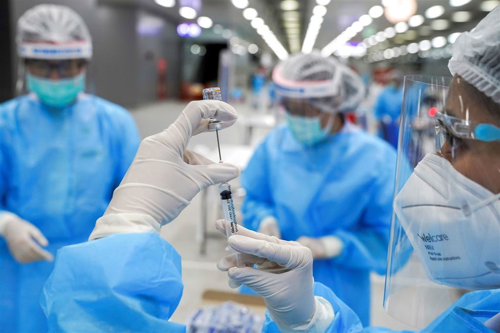 Tre læger står i et vaccinecenter med blå engangskitler, masker, handsker og visir og trækker vacciner op i kanyler.