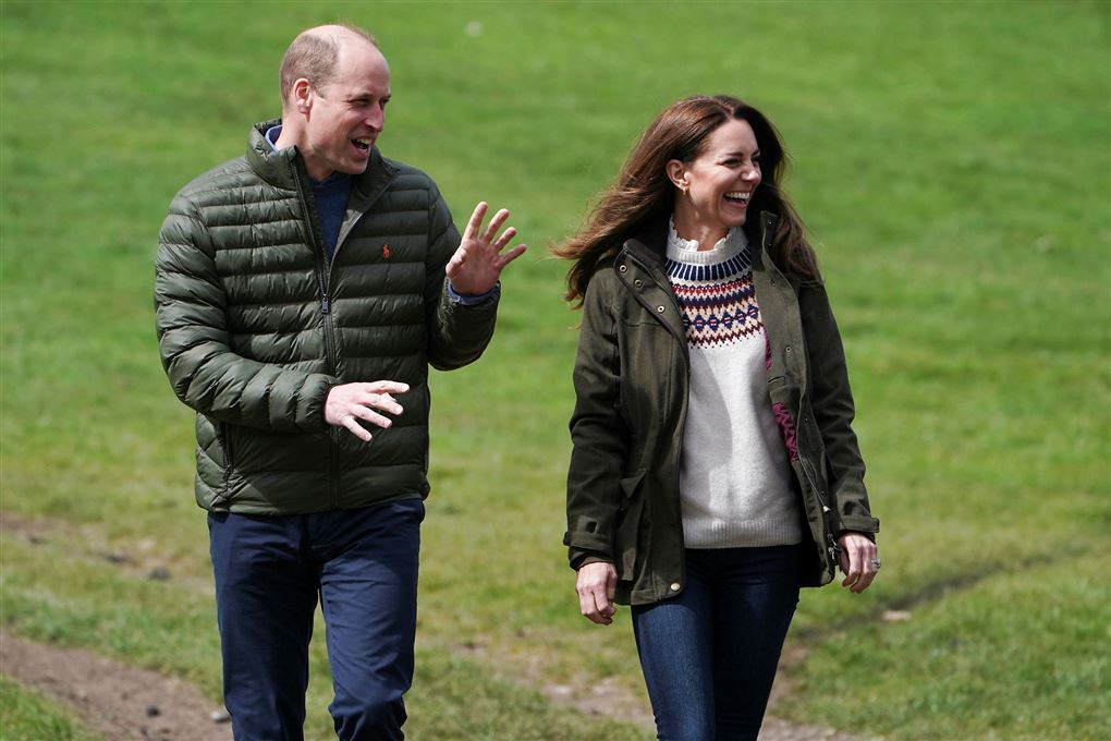 Prins William og Kate i jeans, sweatre og windbreaker jakker på en græsplæne. De griner. 