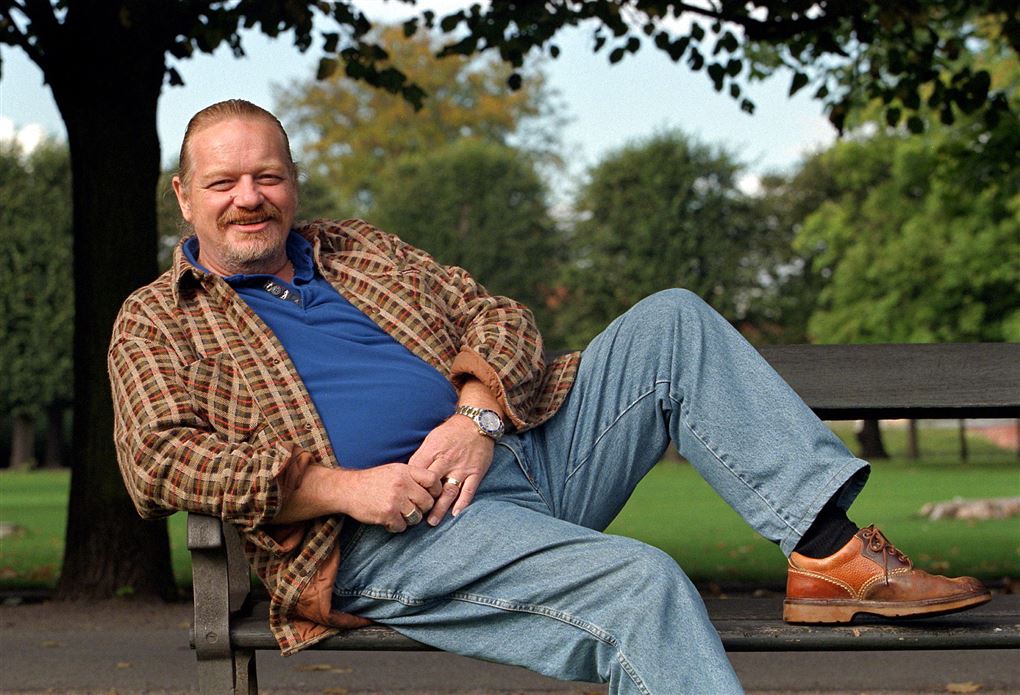 Flemming Bamse Jørgensen sidder henslængt med det ene ben oppe på en bænk i en park. 