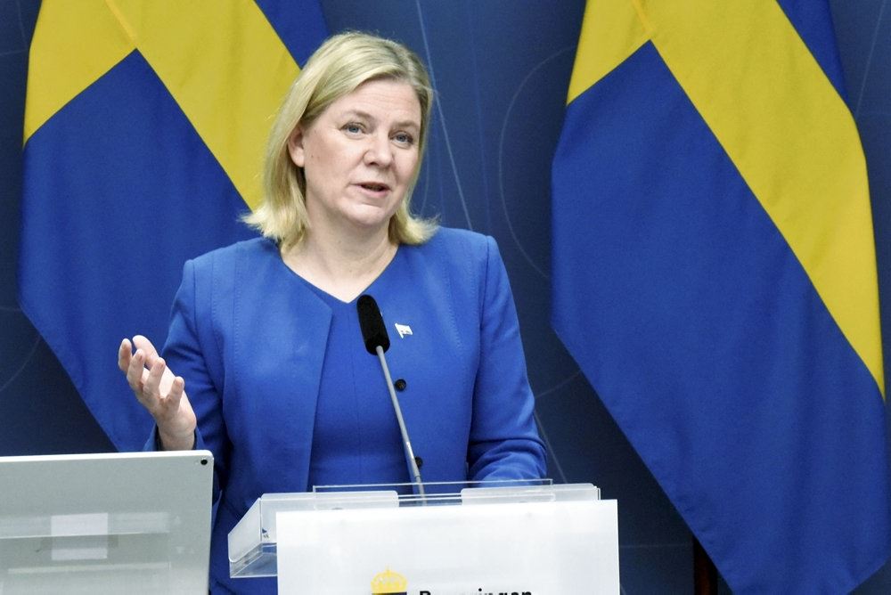 en blond kvinde på talerstol med svensk flag i baggrunden