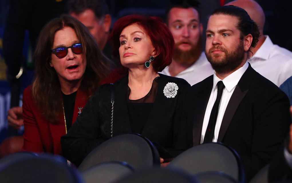 Ozzy Osbourne sidder med solbriller på ved siden af sin kone Sharon, som ser sur ud. Parrets søn Jack Osbourne er også med på billedet. Han sidder ved siden af sin mor med et sort jakkesæt og en hvid skjorte på.
