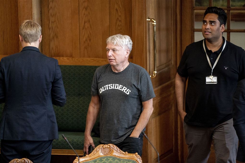 Politiker Uffe Elbæk i Folketingssalen iført mørkegrå t-shirt med hvide bogstaver, hvor der står Outsiders på maven. Bag ham ses Sikandar Siddique der er iført en sort t-shirt. 