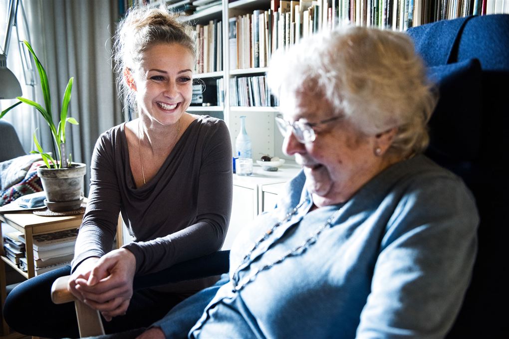 En smilende kvinde sidder inde i stuen hos en ældre dame