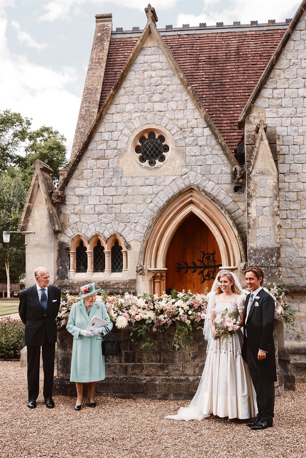 Prins Phillip og dronning Elizabeth står med blomster og kigger hen på prinsesse Beatrice i hvid brudekjole og hendes mand i jaket foran en flot kirkeport