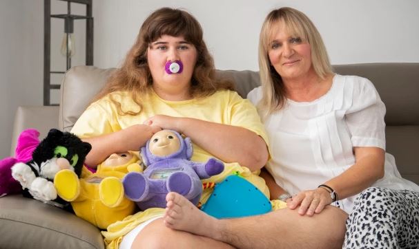 En stor teenager sidder med sin mor i en sofa. Teenageren har en sut i munden og tøjdyr på skødet