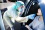 en kvinde i en blå bil bliver corona-testet af en sygeplejerske der er iført engangskittel, engangs handsker, hårbeskyttelse. mundbind og visir. 