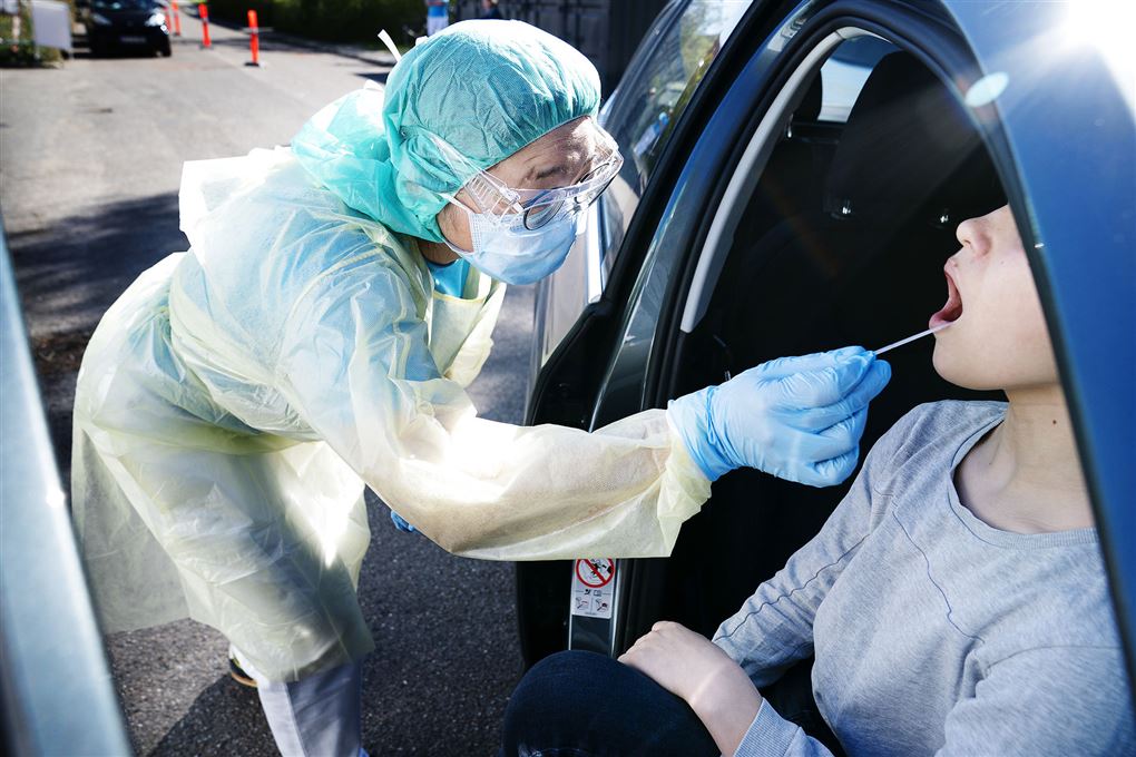 en kvinde i en blå bil bliver corona-testet af en sygeplejerske der er iført engangskittel, engangs handsker, hårbeskyttelse. mundbind og visir. 