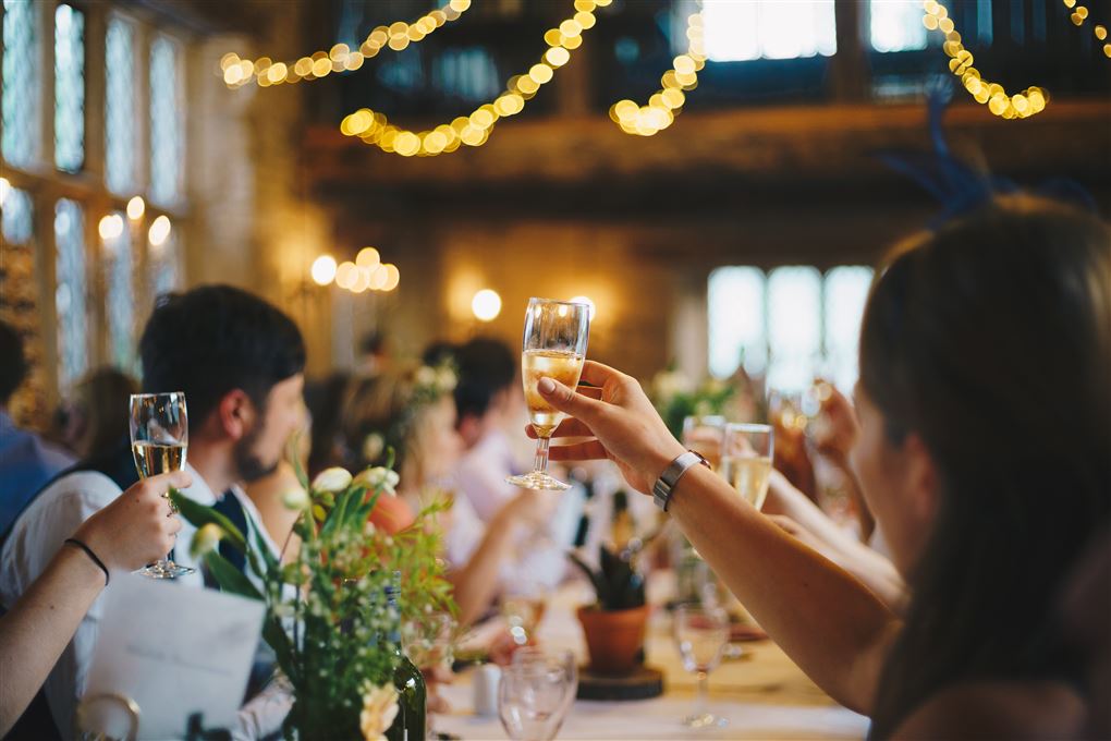 En forsamling skåler i champagneglas efter lyskæderne i loftet og bordpynten at dømme, kunne der være tale om et bryllup.