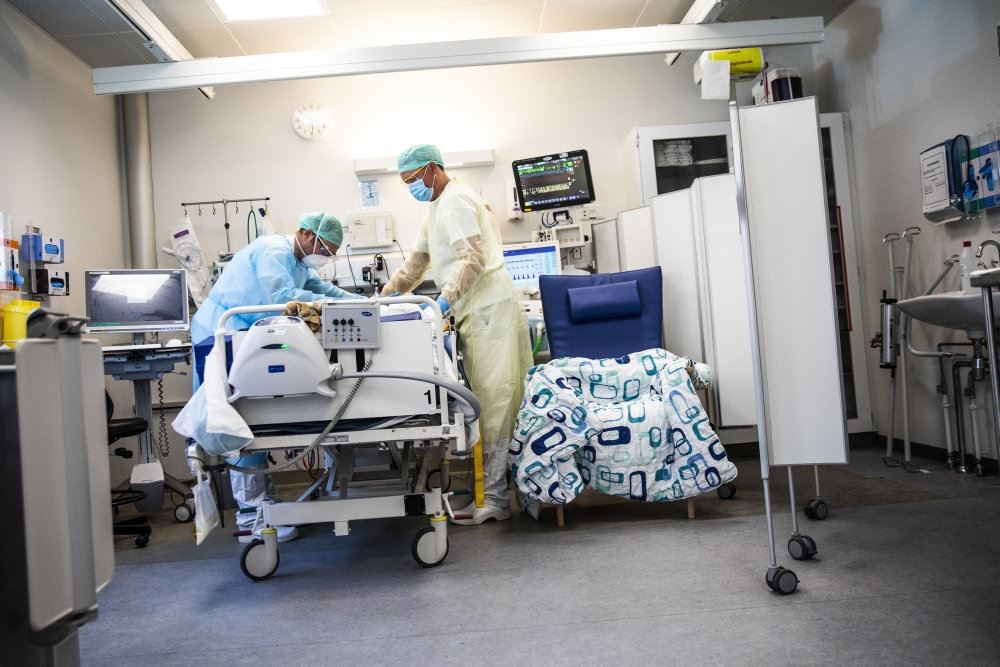 En hospitalsstue, hvor to læger eller sygesplejersker står ved en hospitalsseng med engangskitler, hårnet, handsker og masker på. 