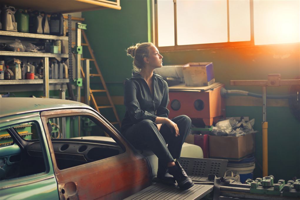En ung kvinde i arbejdstøj sidder på en gammel bil i en garage