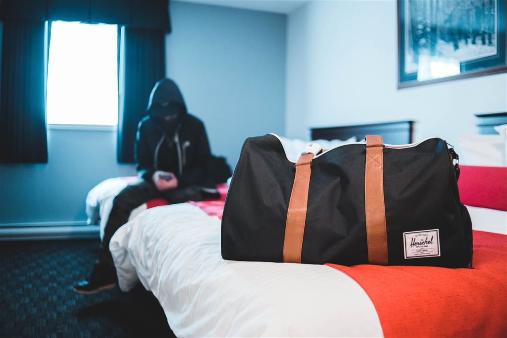 En dreng sidder på en seng med en hætte trukket over ansigtet så man ikke kan se hans identitet. I forgrunden står en taske, der er pakket. 