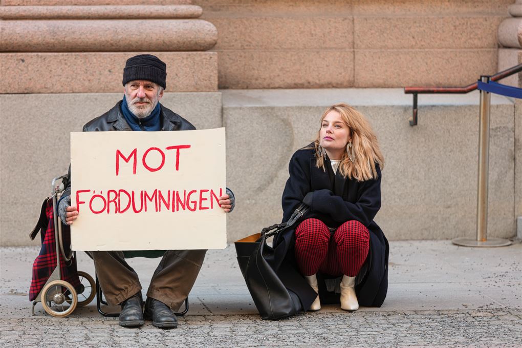 En mand, der ligner en hjemløs sidder med et skilt på gaden ved siden af ham sidder en yngre kvinde med lyst, krøllet hår. På hans skilt står der "Mot fordümningen".