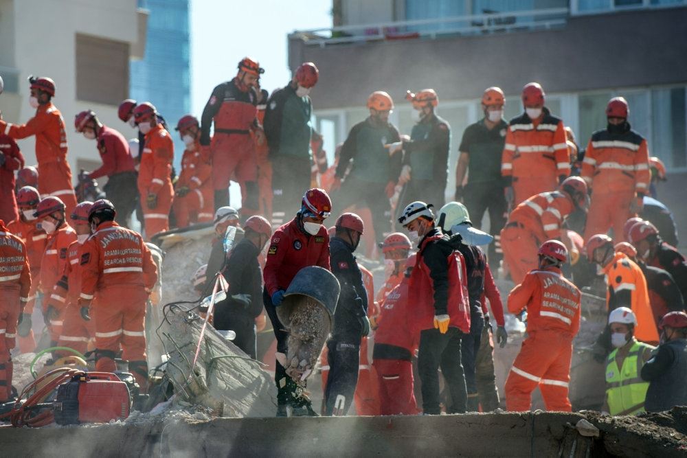 Redningsarbejdere ved murbrokkerne. De er klædt i orange dragter og omgivet af støv. Der er mindst 20 redningsfolk på billedet.