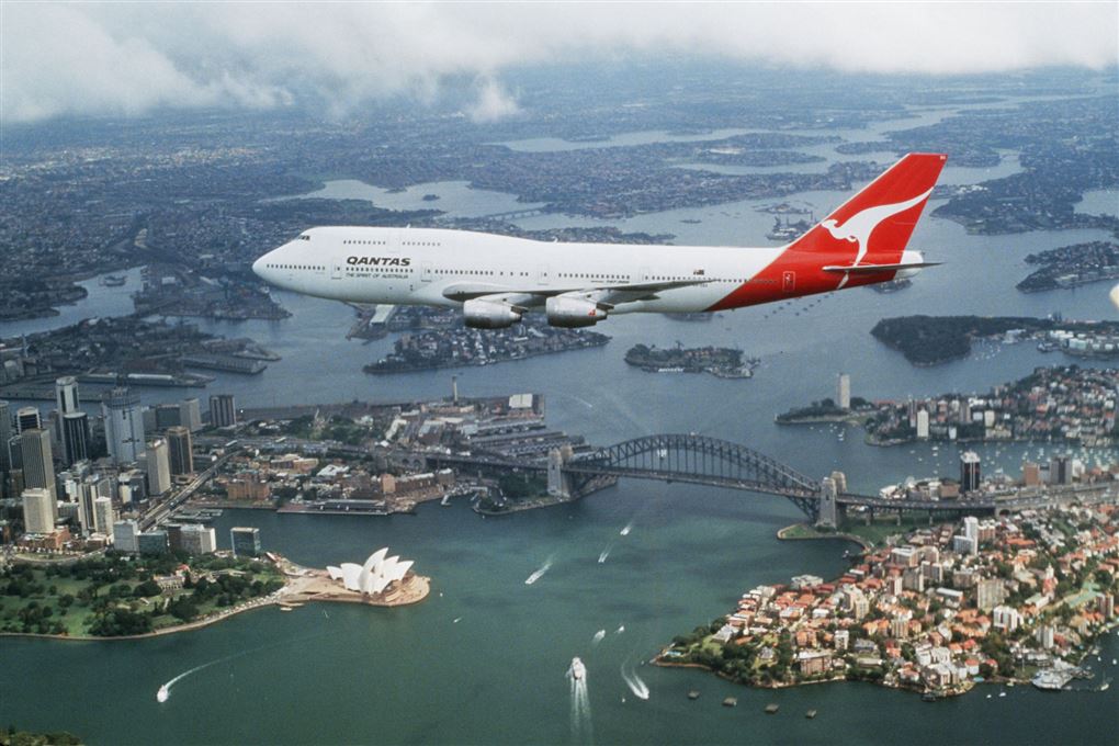 En jumbojet flyver hen over Sydney havn med operahuset og broen nedenunder.