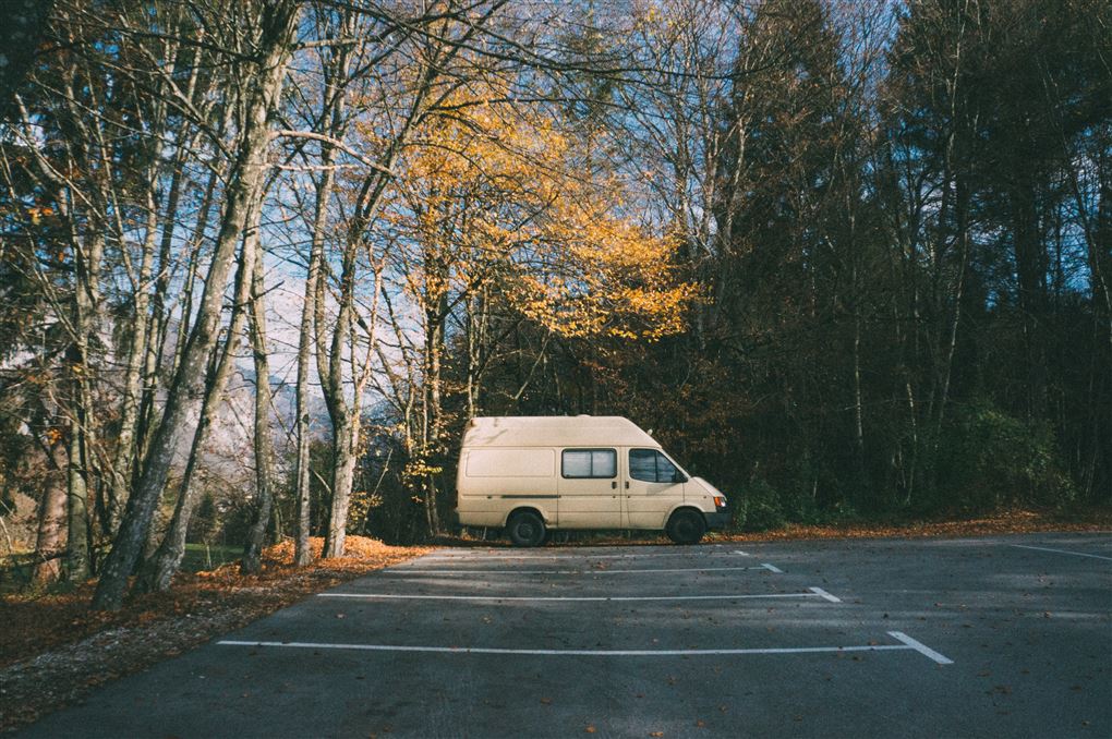 En gammel hvid varevogn parkeret alene på en parkeringsplads med visne træer omkring.
