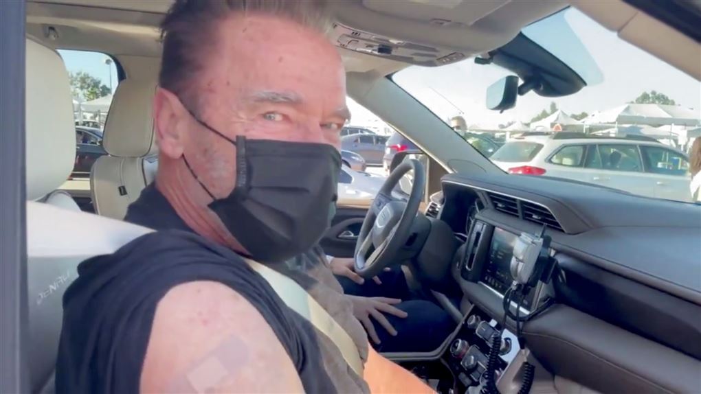 Arnold Schwarzenegger sidder i bil med sort mundbind og plaster på skulderen 