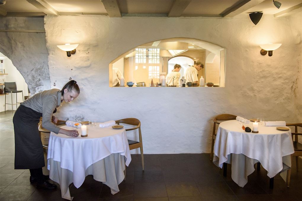 En kvindelig tjener læner sig ind over et bord med en hvid dug og retter på et stearinlys, Bag hende ses to kokke igennem et "hul" i væggen fra restaurant til køkken.