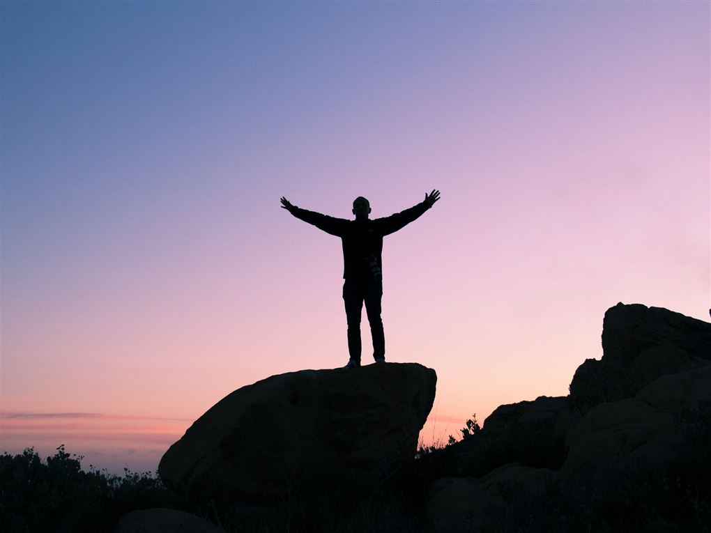 En mand rækker armene i vejret på en klippe i solnedgang