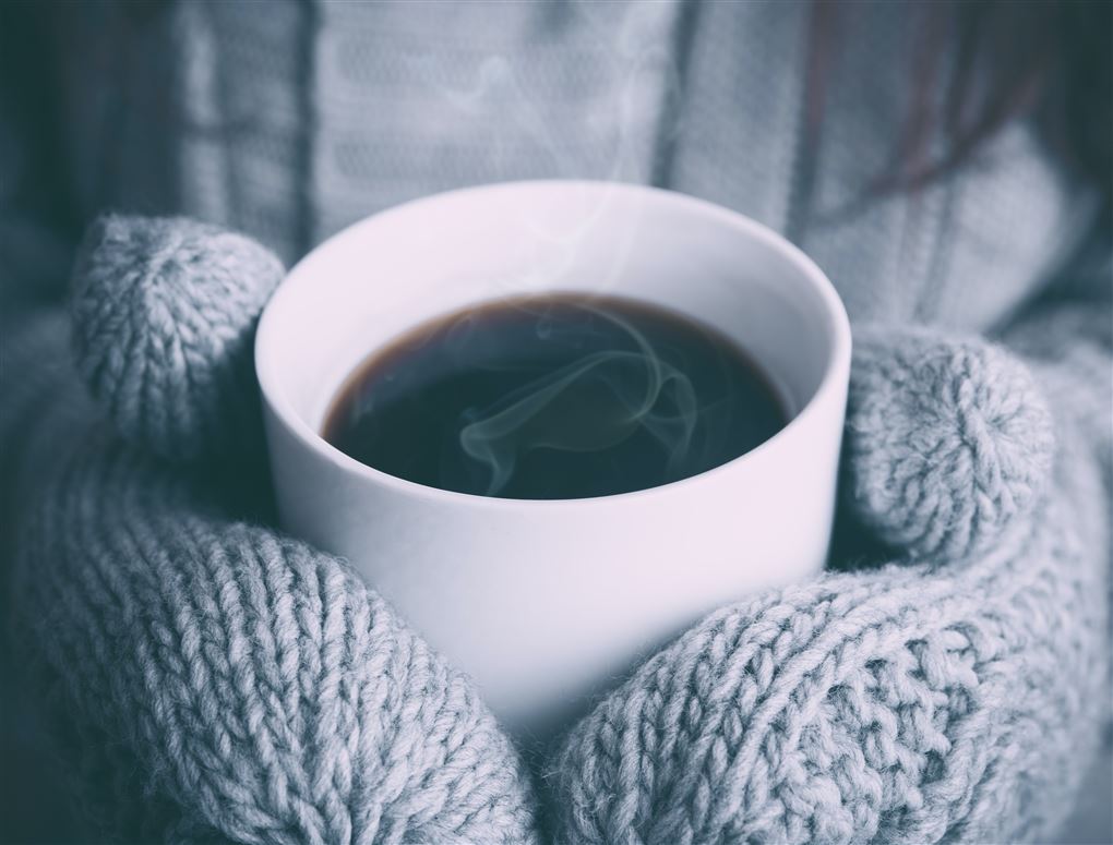 Et par hænder med vanter holder en kop varm kaffe