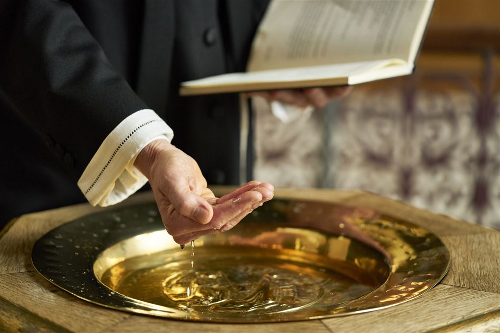 præst med bibel i dene ene hånd og den anden i døbefont