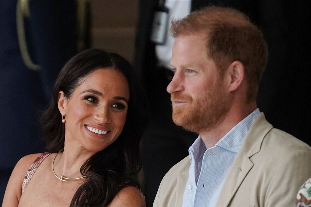 Meghan i sommerkjole læner sig smilende mod Harry i lys jakke og skjorte